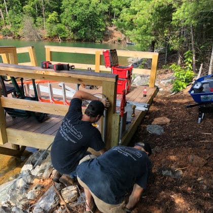 Workers installing a wooden dock by a lake, surrounded by trees and tools.