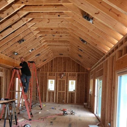 Construction worker on ladder installing lighting in a wooden-framed home interior.