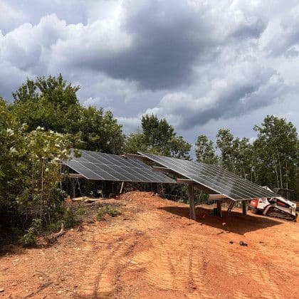 Solar panels installed on a hillside with cloudy skies overhead in a forested area.