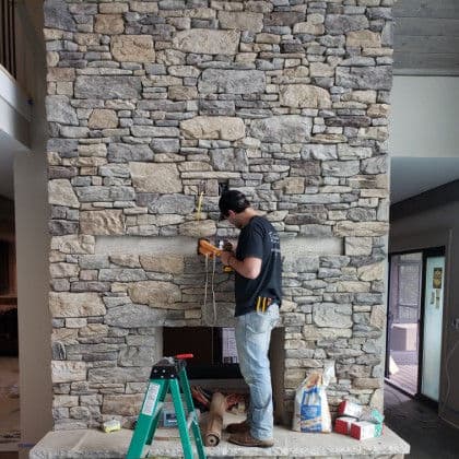 Man installing a wooden mantel on a stone fireplace in a modern home interior.