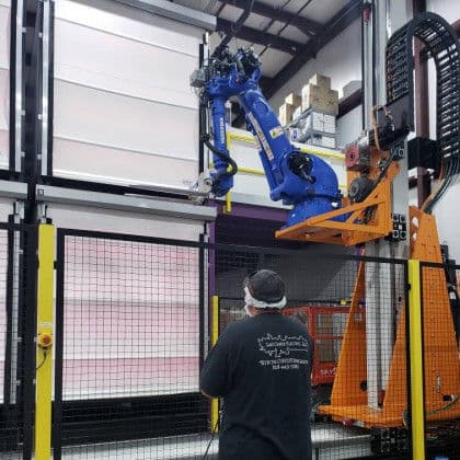 Industrial robotic arm operating in warehouse with technician monitoring process.